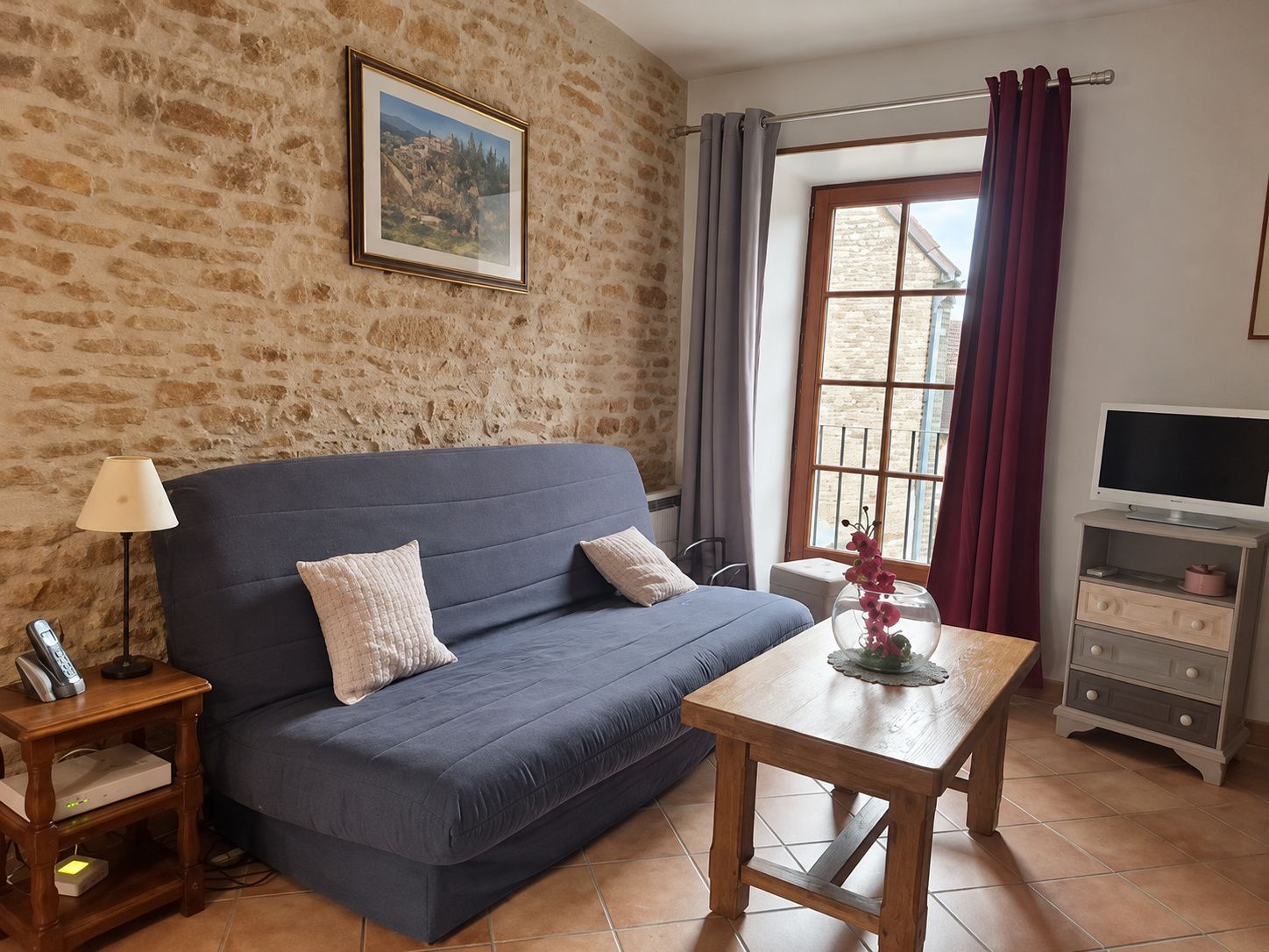 Una habitación con pared de piedra, un sofá azul, una mesa de madera, un televisor sobre un mueble bajo y una ventana con cortinas.