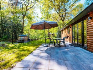 A patio with a table and chairs, an umbrella, and a wooden cabin with large windows. - Holiday Home for Four in Ruinen (Ruinen)