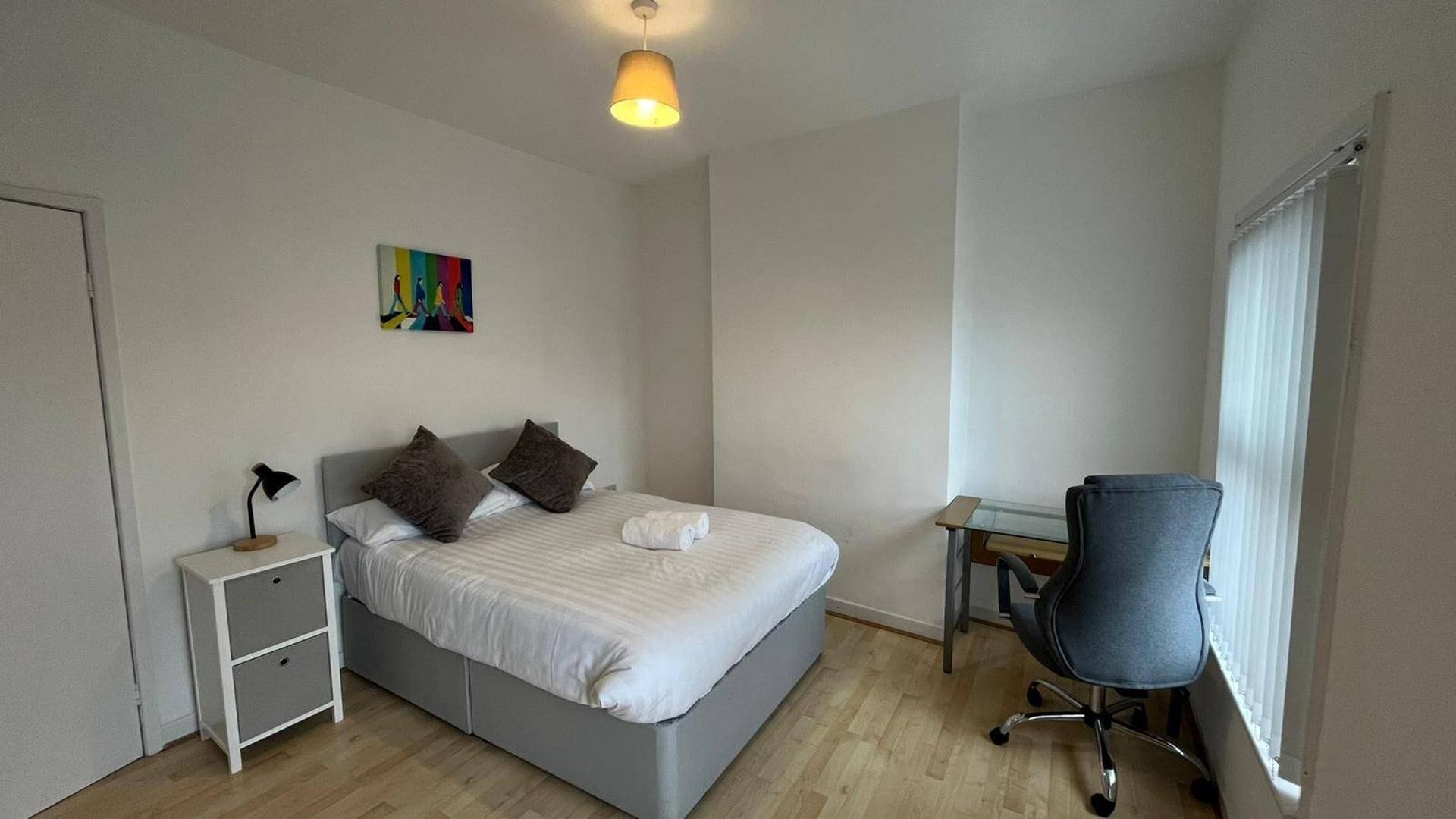 Een slaapkamer met een bed, nachtkastje, lamp, bureau, stoel en een raam met jaloezieën.
