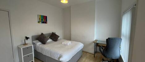 Een slaapkamer met een bed, nachtkastje, lamp, bureau, stoel en een raam met jaloezieën.