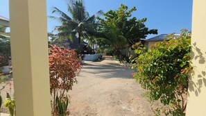 Un sentier menant à un bâtiment dont le toit est recouvert de chaume, entouré d’une végétation tropicale et sous un ciel dégagé.