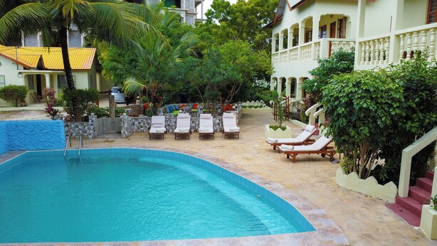 A swimming pool with lounge chairs, surrounded by a landscaped garden and a building with a balcony. - Bustani Oasis with AC,Pool, Family-Friendly, Couples, Beach 5 Min Walk (Nungwi)