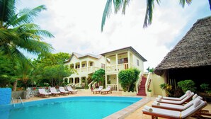 A swimming pool with lounge chairs, a building with balconies, and a thatched-roof structure. - Bustani Oasis with AC,Pool, Family-Friendly, Couples, Beach 5 Min Walk (Nungwi)