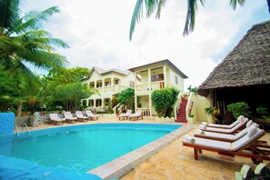 A swimming pool with lounge chairs, a building with balconies, and a thatched-roof structure. - Bustani Oasis with AC,Pool, Family-Friendly, Couples, Beach 5 Min Walk (Nungwi)