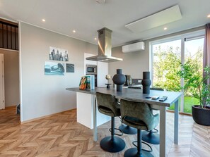 A modern kitchen with a central island, a dining area with bar stools, and a large window offering a view of greenery. - Modern Waterfront Villa Retreat for 4 (Wanneperveen)