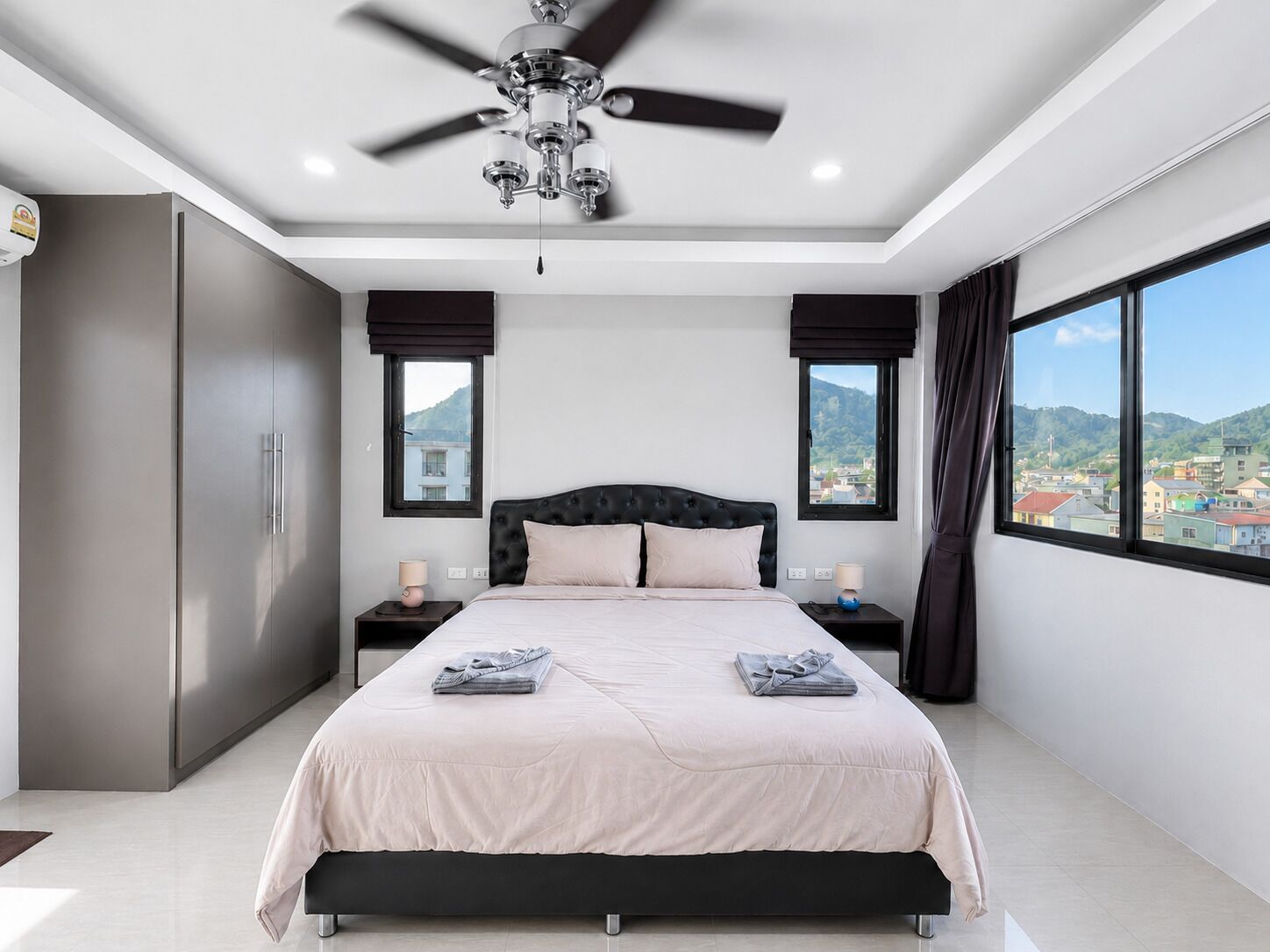 Un dormitorio moderno con una cama grande, dos mesitas de noche, un ventilador de techo y vistas a un paisaje urbano a través de amplias ventanas.