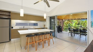 A modern kitchen with a central island, bar stools, and a view of an outdoor dining area. - Tide n Town - 2 bedroom resort apartment - pools, restaurant (Agnes Water)