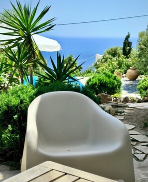 A patio with a white plastic chair, a white umbrella, and a view of the sea. - Studio with magnificent sea view and gardens (Kastri)