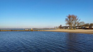 Ruhiges Gewässer mit blauem Himmel, einem Sandstrand und einer Reihe von Häusern im Hintergrund.