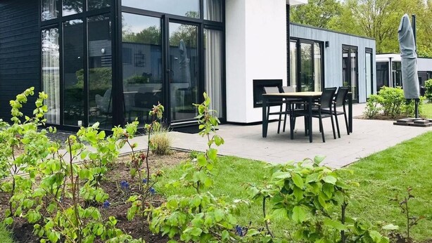 Ein modernes Haus mit großen Glasfenstern, einer Terrasse mit Tisch und Stühlen und einem Garten mit Pflanzen.