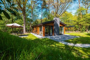 A modern log cabin with a stone chimney, surrounded by lush greenery and a patio with outdoor seating. - 3-bedroom Forest Lodge (Ruinen)