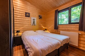 A wooden cabin bedroom with a bed, a nightstand, a wall-mounted lamp, and a window with a view of trees. - 3-bedroom Forest Lodge (Ruinen)