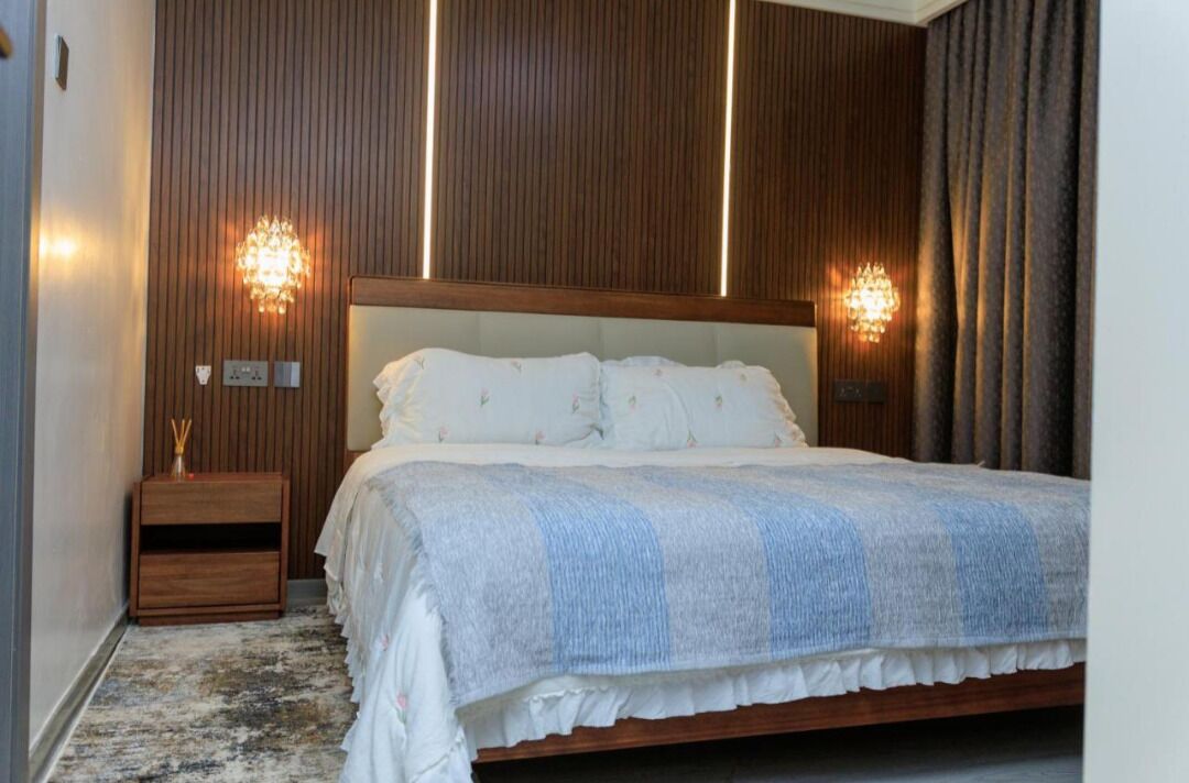 A bedroom with a wooden headboard, a large bed with white bedding, two wall-mounted lights, and a nightstand with a small lamp.