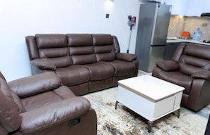 A living room with a brown leather sofa, a white coffee table, and a kitchen area in the background. - Charming apartment in an enchanting Nairobi neighborhood perfect for your stay (Nairobi)
