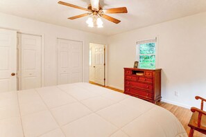 A bedroom with a large bed, a wooden dresser, a ceiling fan, and a window with a view of greenery. - Creekside Cottage: Kayak, Tube, Fish, Remote Work (Dugspur)