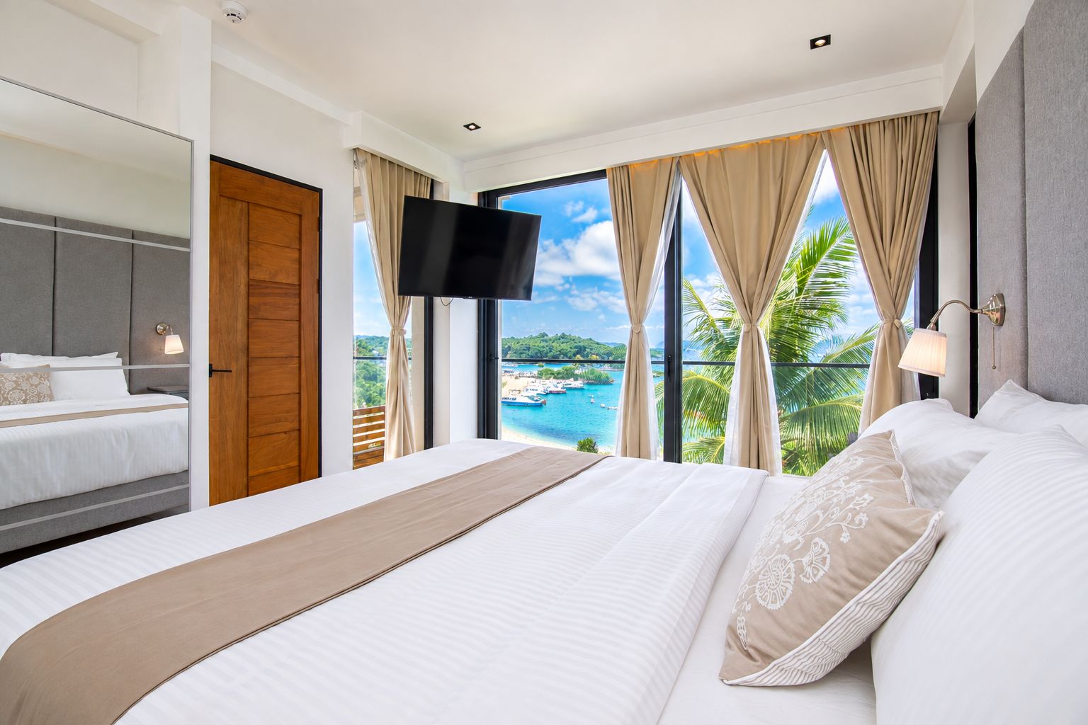A hotel room with a large bed, a television, a balcony with a view of the sea, and a wooden door.