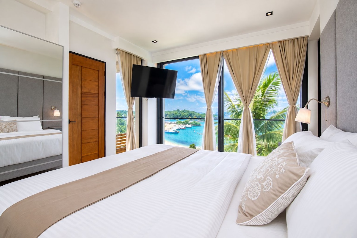Habitación de hotel con una cama grande, televisión, balcón con vista al mar y puerta de madera.
