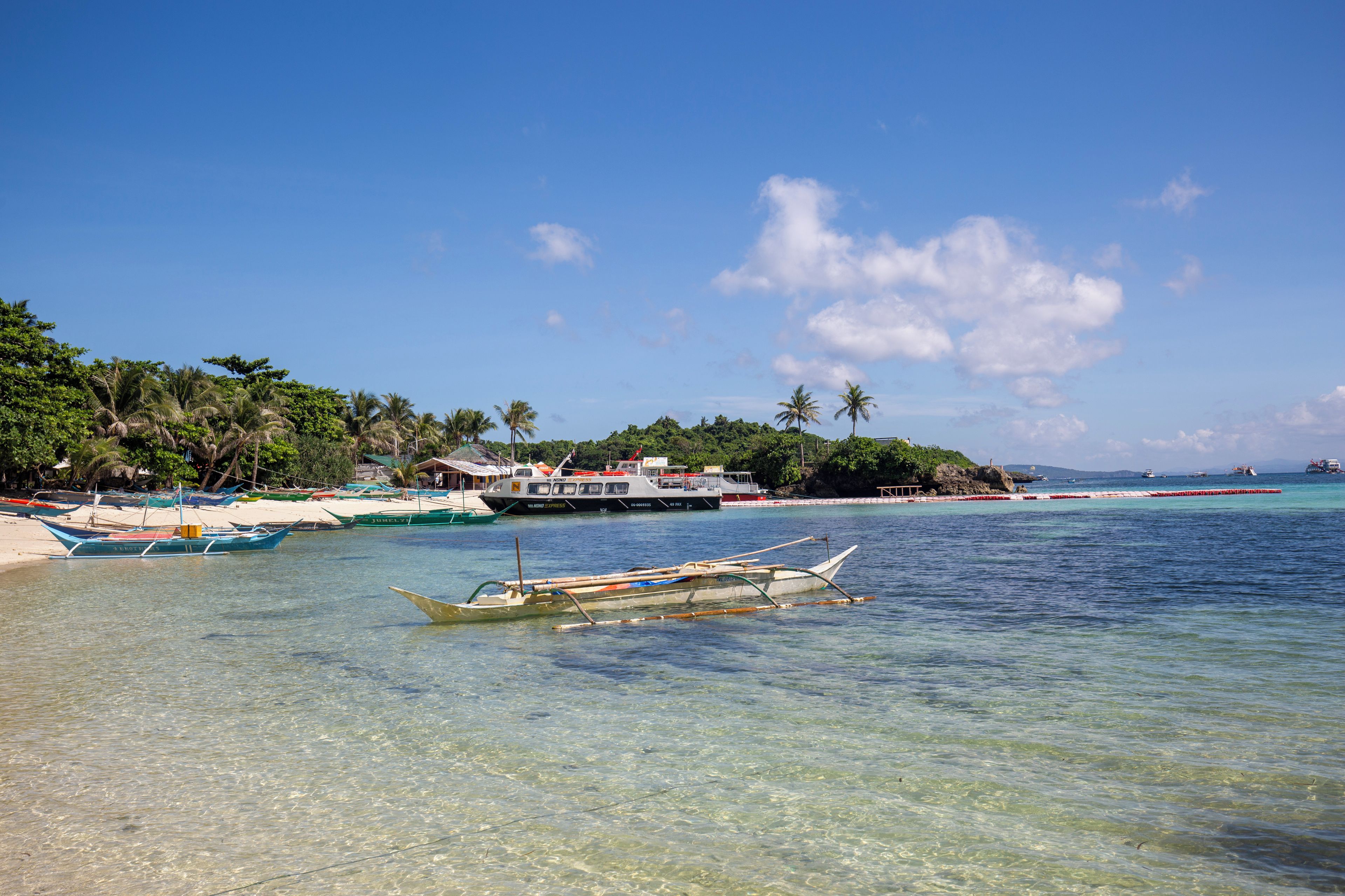 A beach with clear water, boats, and a building with a red roof.