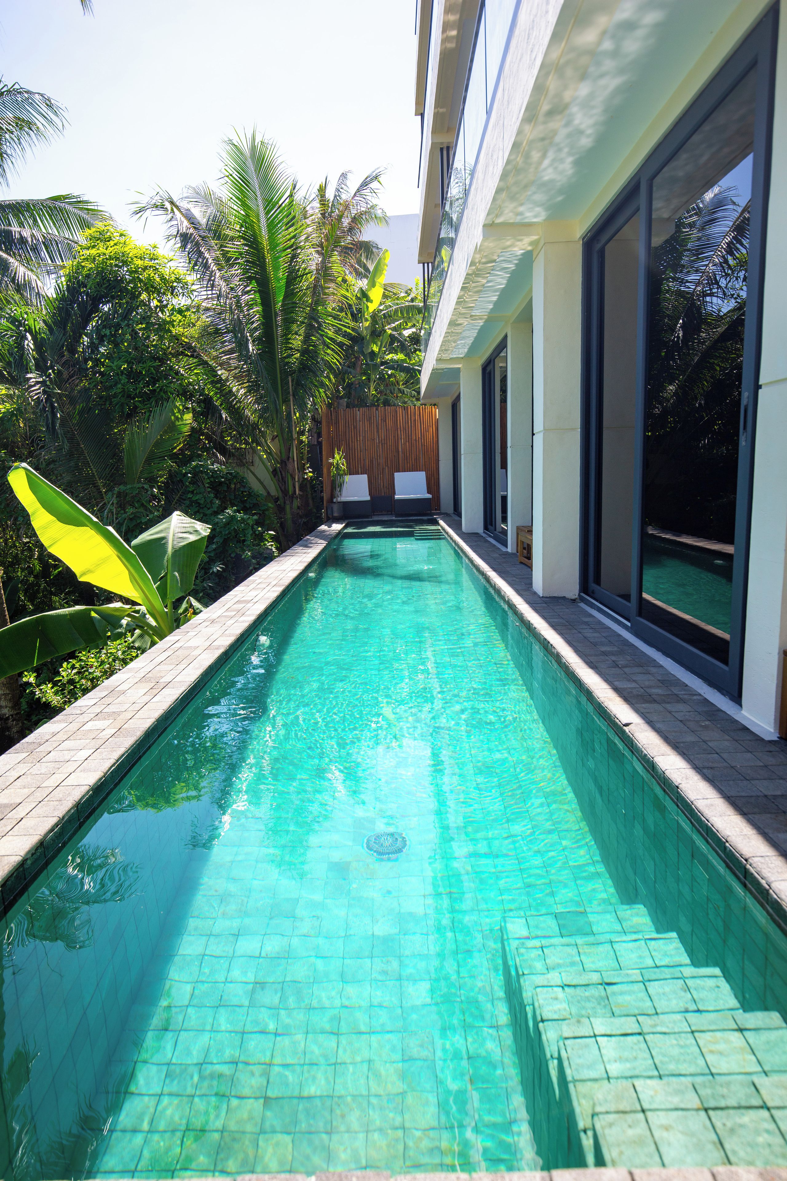 A rectangular swimming pool with clear water, surrounded by lush greenery and a wooden fence.