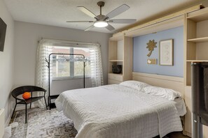 Ein Schlafzimmer mit einem Bett, einem Deckenventilator, einem Fenster mit Vorhängen, einem Stuhl und einer Wand mit einem dekorativen Gegenstand.