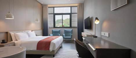 A modern hotel room with a bed, desk, chairs, a TV, and a large window with a view.