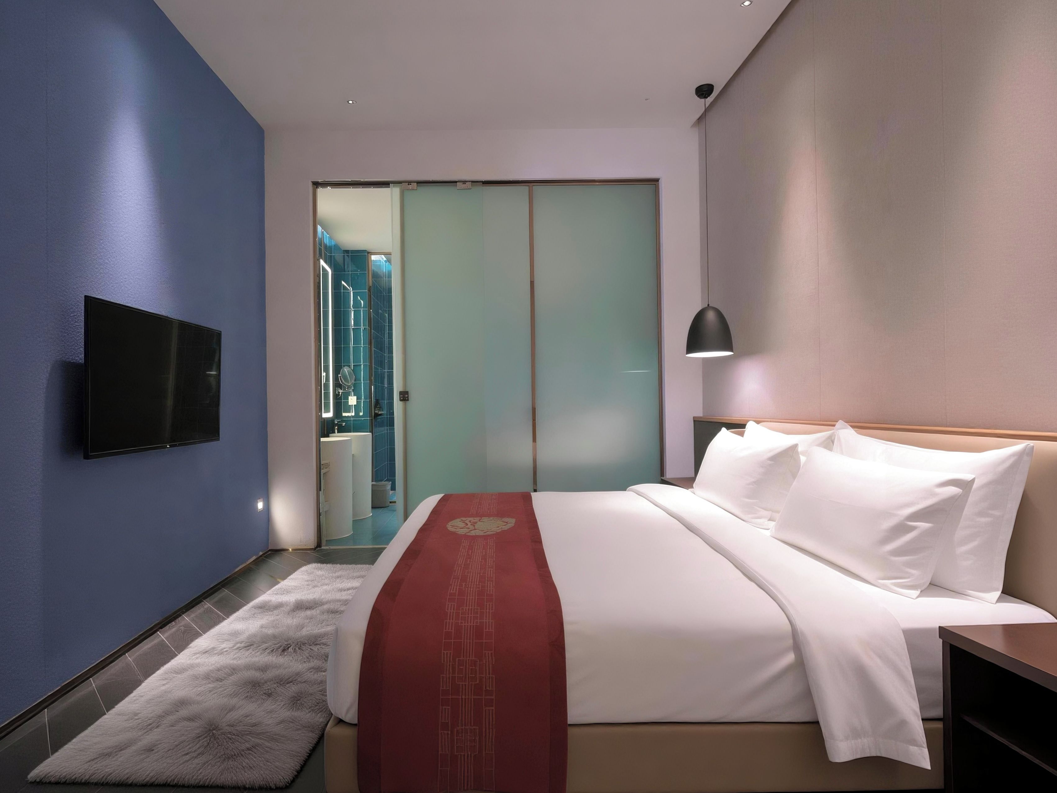 A hotel room with a bed, a TV mounted on the wall, a bedside lamp, and a view of the bathroom through a sliding door.