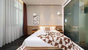 Habitación de hotel con una cama grande, una mesita de noche, una lámpara de pared, un separador de vidrio y una ventana con cortinas.