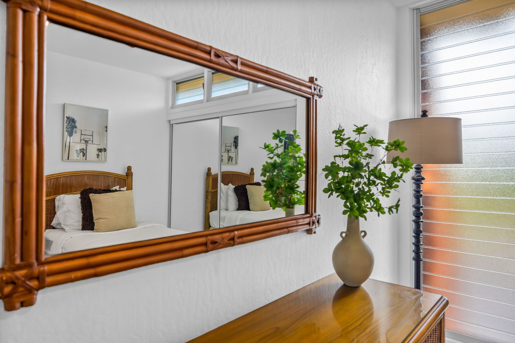 A room with a bed, a mirror, a vase with a plant, and a lamp.