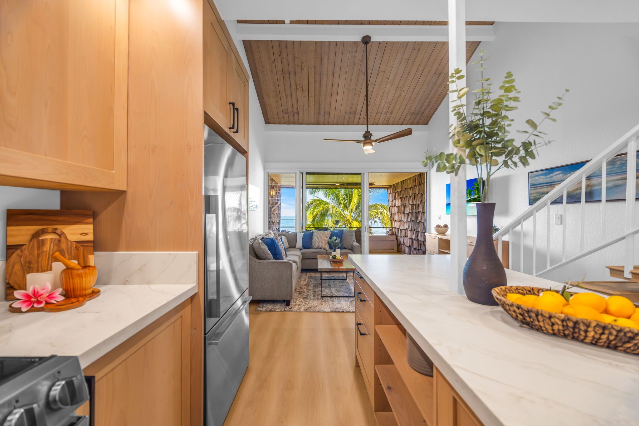 A modern kitchen with wooden cabinets, a large island, and a view of the living area with a sofa and a plant.