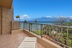 A balcony with a view of the ocean and mountains, a chair, and a stone wall. - Beachfront 2BD w Stunning Ocean View-paki Maui 424 (Kaanapali)