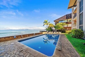 A swimming pool with a view of the ocean, surrounded by a stone deck and a grassy area with palm trees. - 2BD Oceanfront Condo With Pool Near Beach-nohonani 105 (Napili-Honokowai)