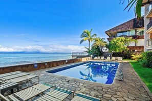 A swimming pool with lounge chairs, a view of the ocean, and a building with a modern design. - 2BD Oceanfront Condo With Pool Near Beach-nohonani 105 (Napili-Honokowai)