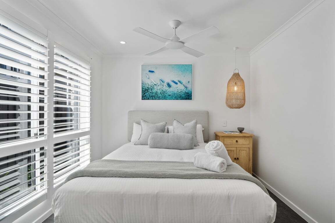 A hotel room with a bed, a ceiling fan, a wall-mounted artwork, a bedside table, and a window with blinds.