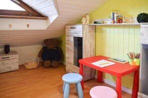 Ein Kinderzimmer mit Holzboden, einem roten Tisch, einem blauen Hocker, einem Teddybären und einem Bücherregal.