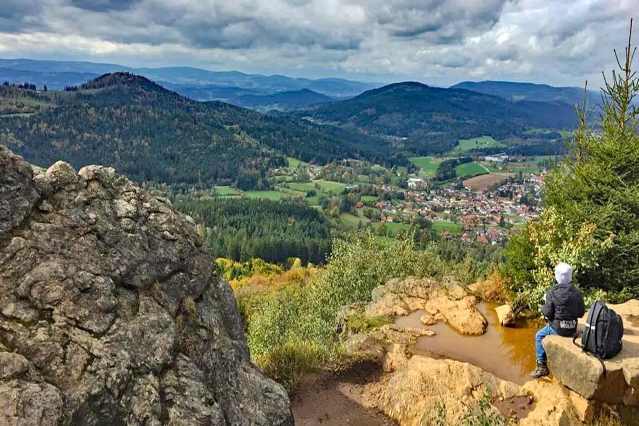 Une personne assise sur un rocher, un sac à dos sur le dos, et qui surplombe une vallée parsemée de maisons et de verdure.