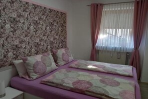 Een slaapkamer met bloemenbehang, een bed met een geborduurd sprei en roze gordijnen.
