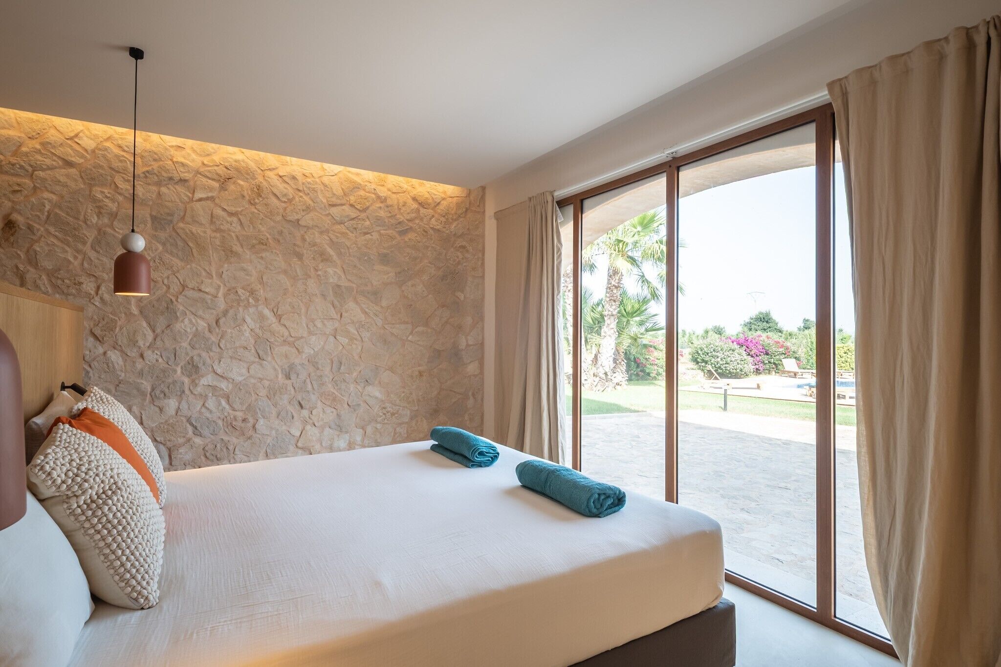 Een slaapkamer met een stenen muur, een bed met twee kussens, een hanglamp en uitzicht op het omliggende landschap.