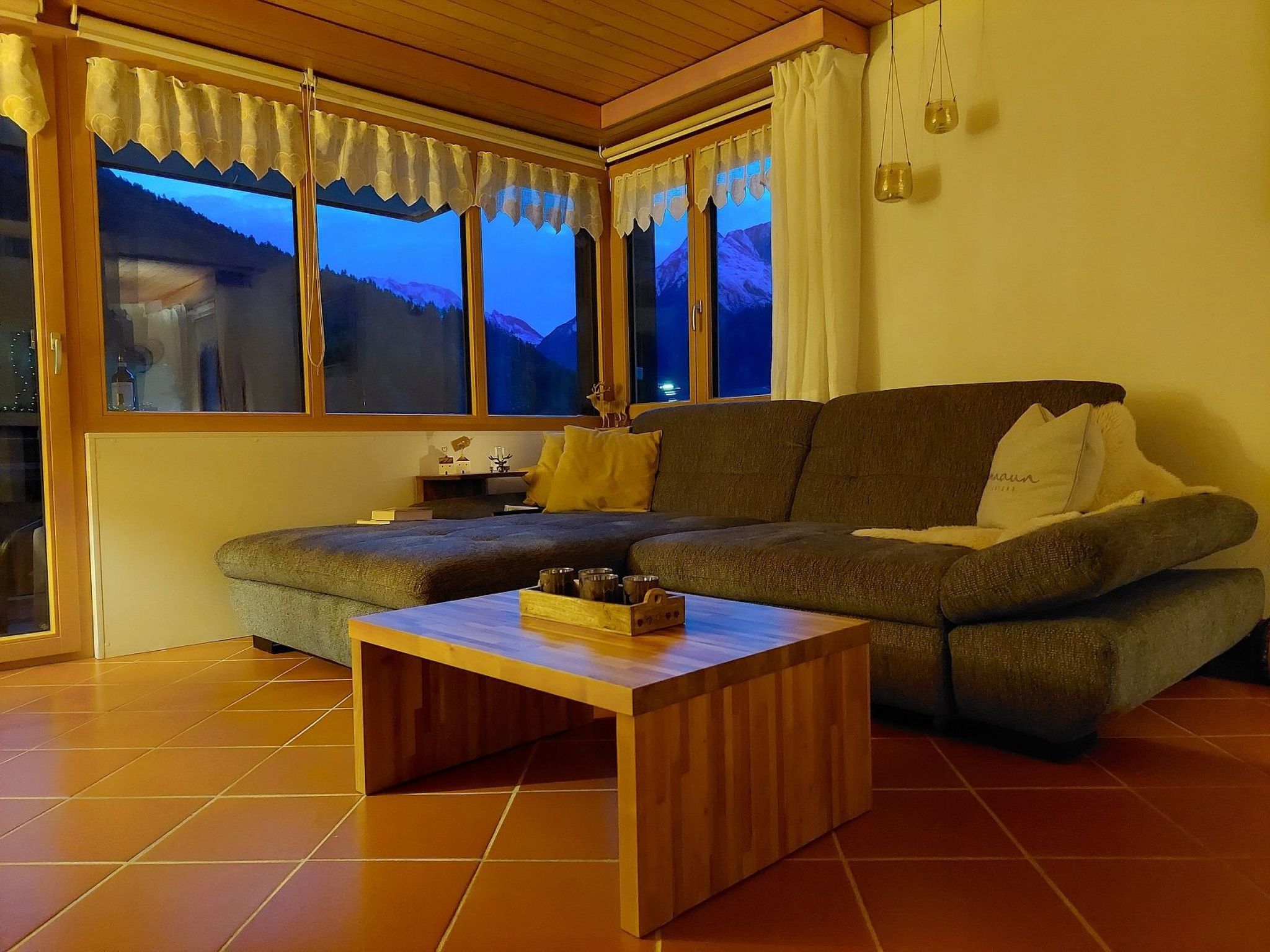 A cozy living room with a sectional sofa, a wooden coffee table, and large windows with curtains.
