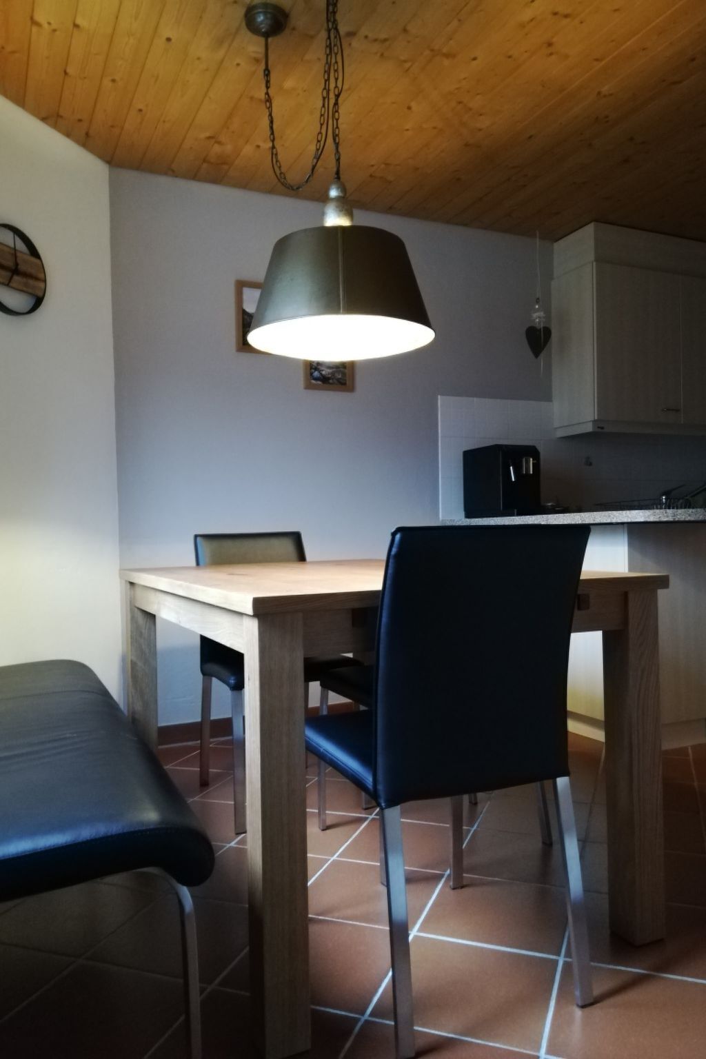 A dining area with a wooden table and chairs, a hanging light fixture, and a kitchen in the background.