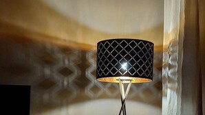 Eine Stehlampe mit geometrischem Lampenschirm, ein kleiner Topfpflanze und ein Magazinhalt auf einem Ständer, in einem Zimmer mit Holzbalkendecke und schlichter Wand.