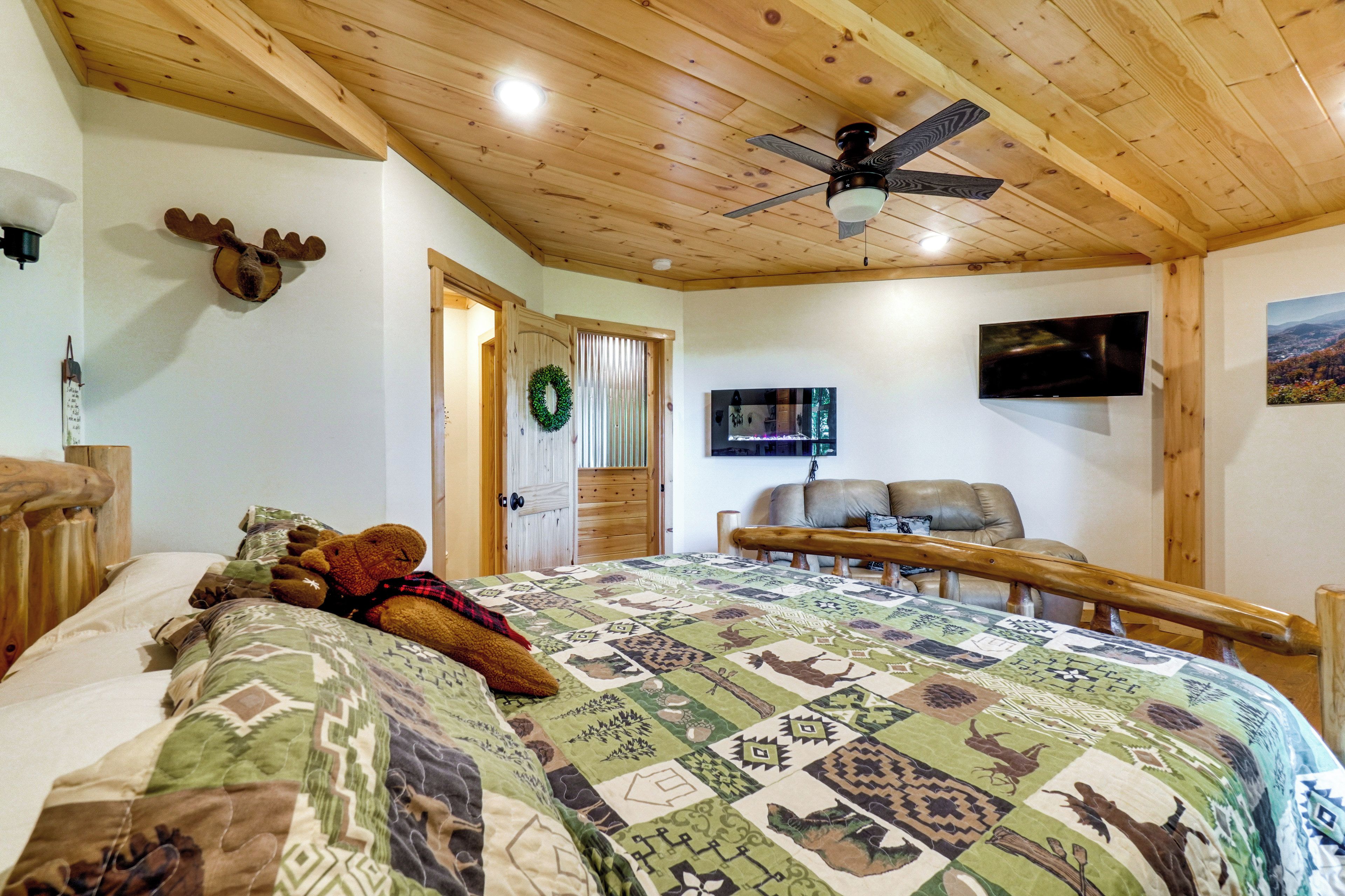 A bedroom with a wooden ceiling, a bed with a patterned quilt, a mounted television, and a moose head decoration.