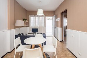 Hotellihuone, jossa on ruokailutila, jossa on pyöreä pöytä ja valkoiset tuolit, televisio ja ikkuna, jossa on verhot.