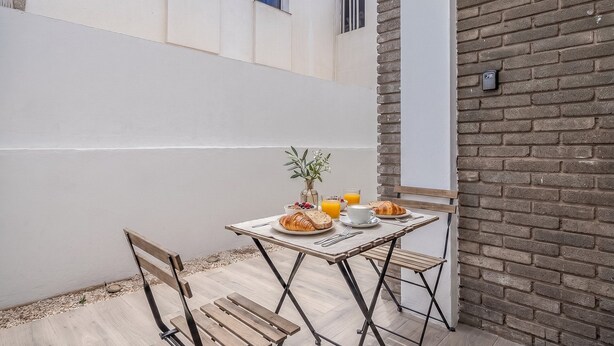 Un petit espace extérieur aménagé pour prendre les repas, avec une table pour deux, un banc et un mur en briques.