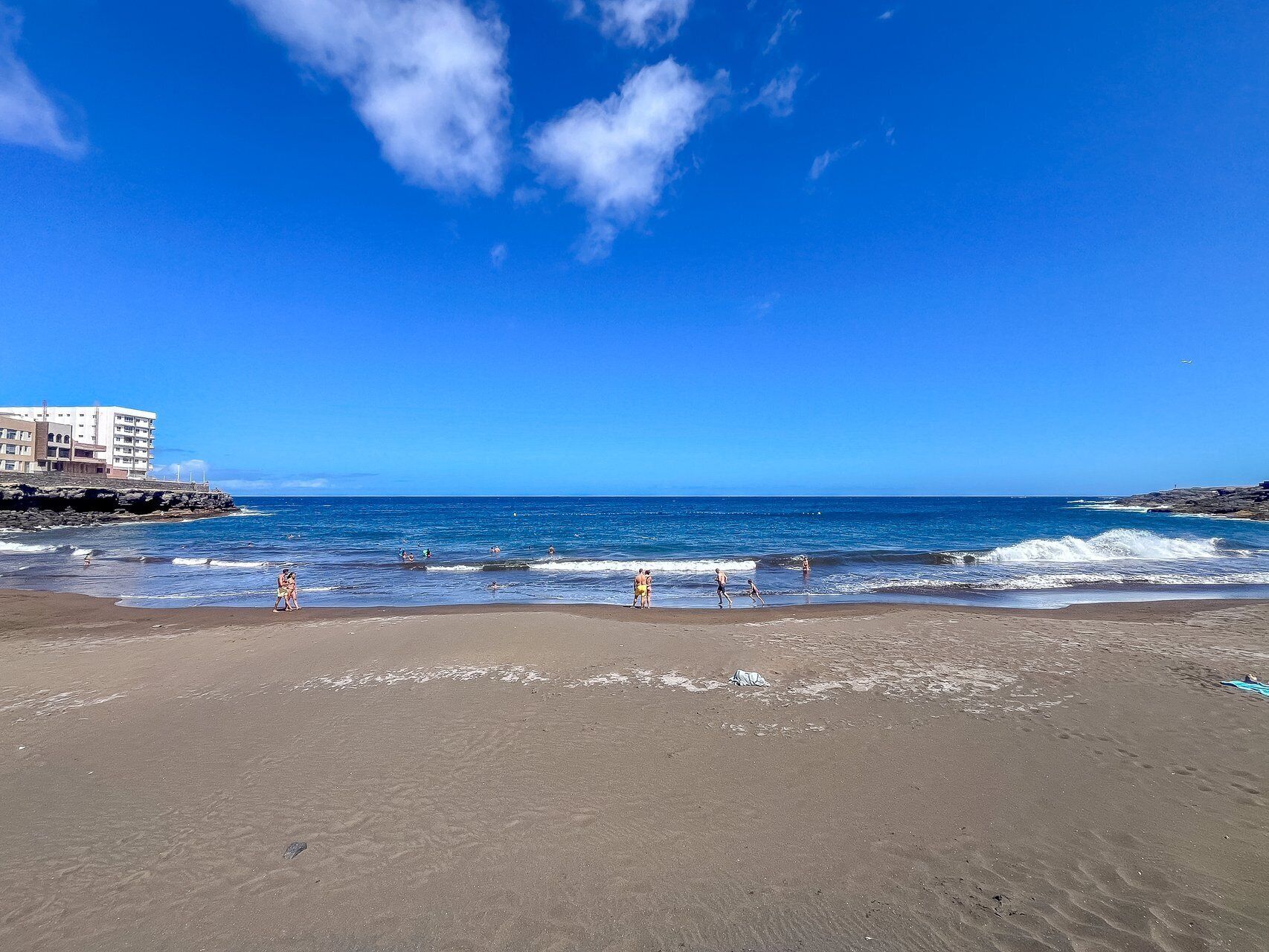 Une plage avec un bâtiment en arrière-plan, des vagues et des gens.