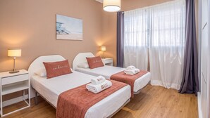 Une chambre d’hôtel avec deux lits, un tableau représentant une plage, une fenêtre avec des rideaux et une lampe sur chaque table de chevet.