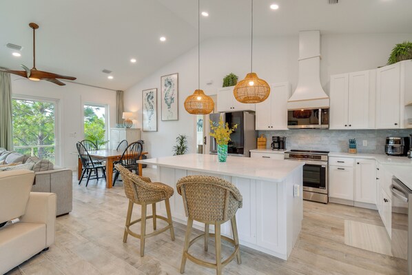 A modern kitchen with white cabinetry, a central island with bar stools, pendant lighting, and a dining area with a table and chairs. - Fort Morgan Hideaway Hot Tub | Golf | Beach Access (Gulf Shores)