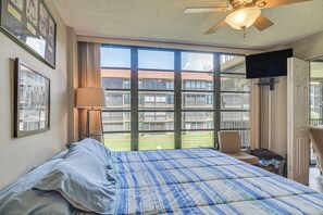 Ett hotellrum med en stor säng, en takfläkt, ett fönster med utsikt över en byggnad och en tv som är monterad på väggen.