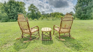 Deux fauteuils à bascule et une petite table sont placés dans un champ herbeux, avec des arbres en arrière-plan.