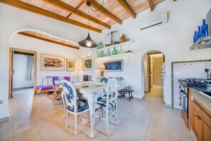 A dining area with a white table and chairs, a hanging light fixture, and a kitchen with a stove and cabinets. - Romantic Country Hideaway in Pollensa (Pollença)