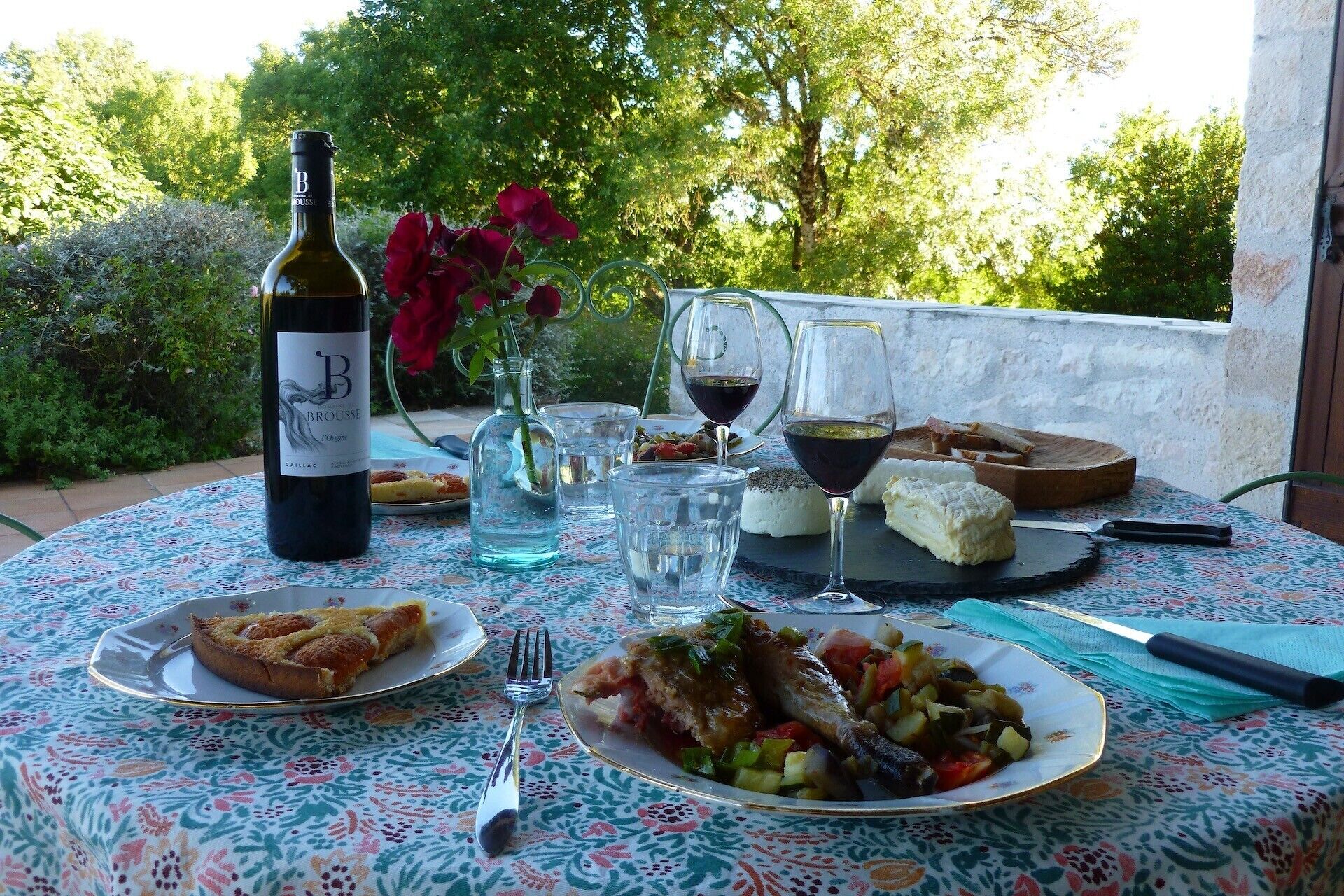 Een tafel buiten gedekt met een fles wijn, glazen, een bord eten en een vaas vol bloemen.